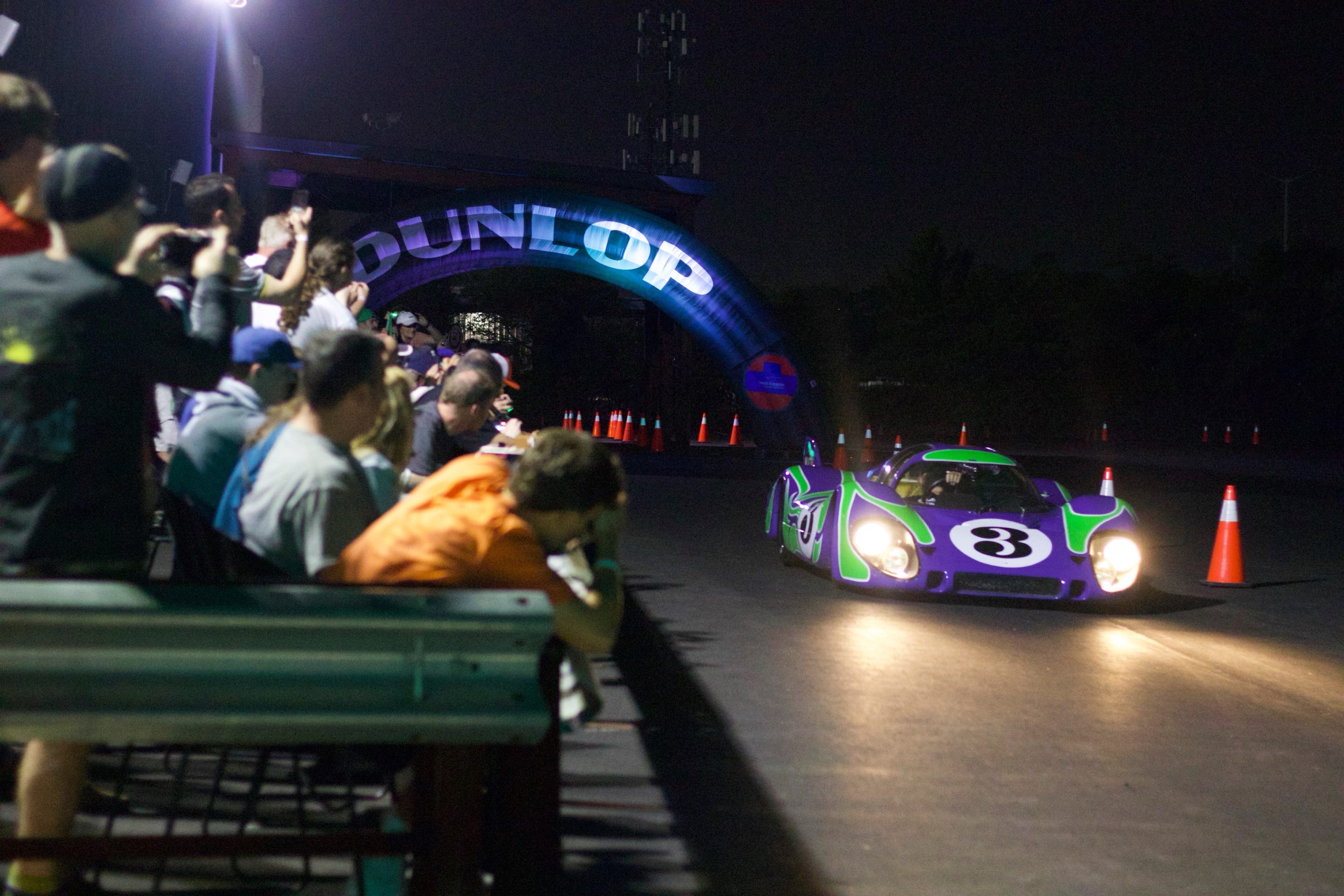 A Porsche 917 with the famous hippie livery driving under a mini Dunlop Bridge and past traffic cones in front of a grandstand of fans